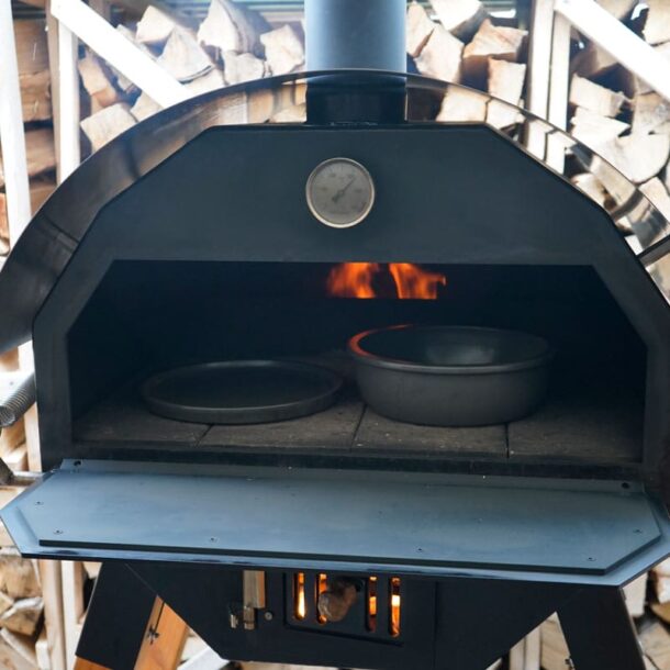 Holzbackofen offen: Merklinger-Holzbackofen mit geöffneter Klappe; innen zwei runde Keramik-Backformen, hinten sichtbare Flammen; im Hintergrund gestapeltes Brennholz.
