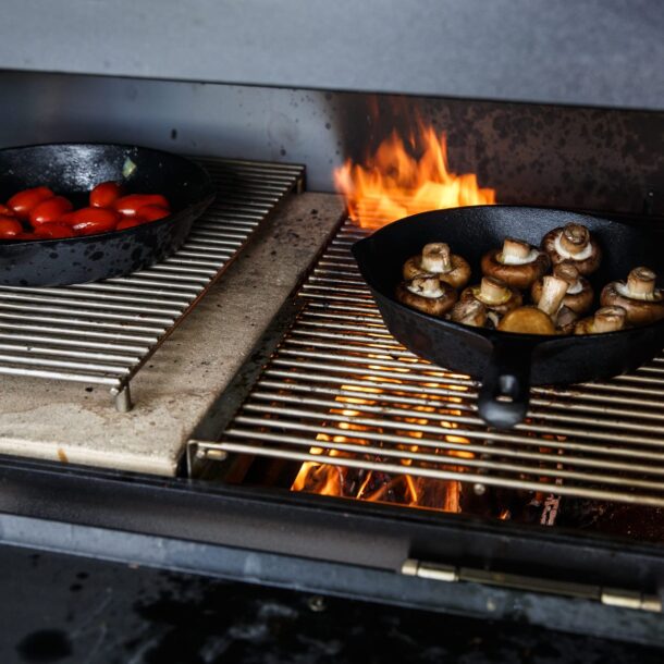 Zwei gusseiserne Pfannen mit Champignons und Kirschtomaten auf doppeltem Grillrost.“ Zwei gusseiserne Pfannen mit Champignons und Kirschtomaten auf doppeltem Grillrost.“