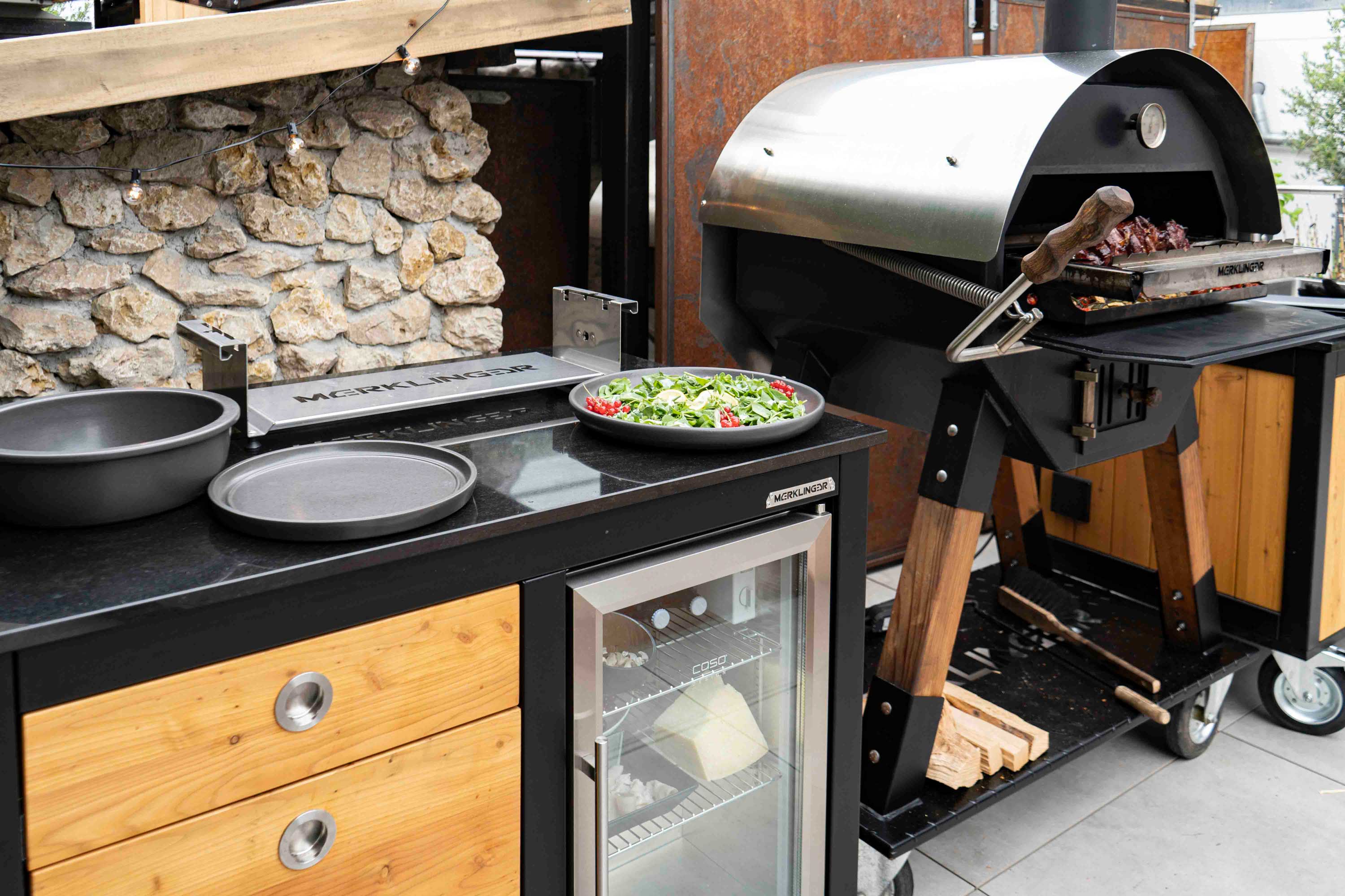 Merklinger Outdoor-Küchenmodul mit Glastür-Kühlschrank und Pizza-Zubehör neben MERKLINGER Holzofen beim Grillen