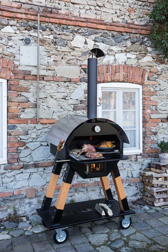 Mobiler Rund-Ofen vor Steinmauer Mobiler MERKLINGER Rundofen auf Rollen vor einer rustikalen Steinmauer Merklinger Holzbackofen Outdoor mit Holzfeuer – Grillen und Backen vor rustikaler Natursteinwand, gefüllt mit Fleisch und Beilagen