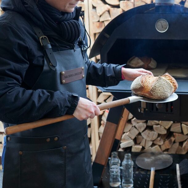 Sicheres Arbeiten am Holzbackofen“ Person mit Leder-Schürze entnimmt mit MERKLINGER Brotschieber ein frisch gebackenes Brot.“