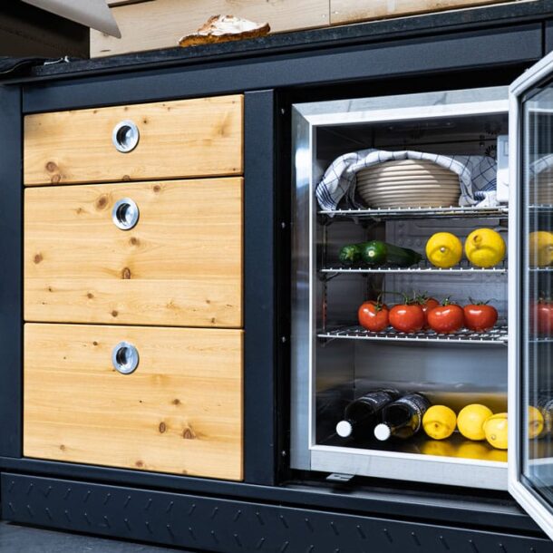 Holz-Küchenmodul mit drei Schubladen links und Glastür-Kühlschrank rechts.“