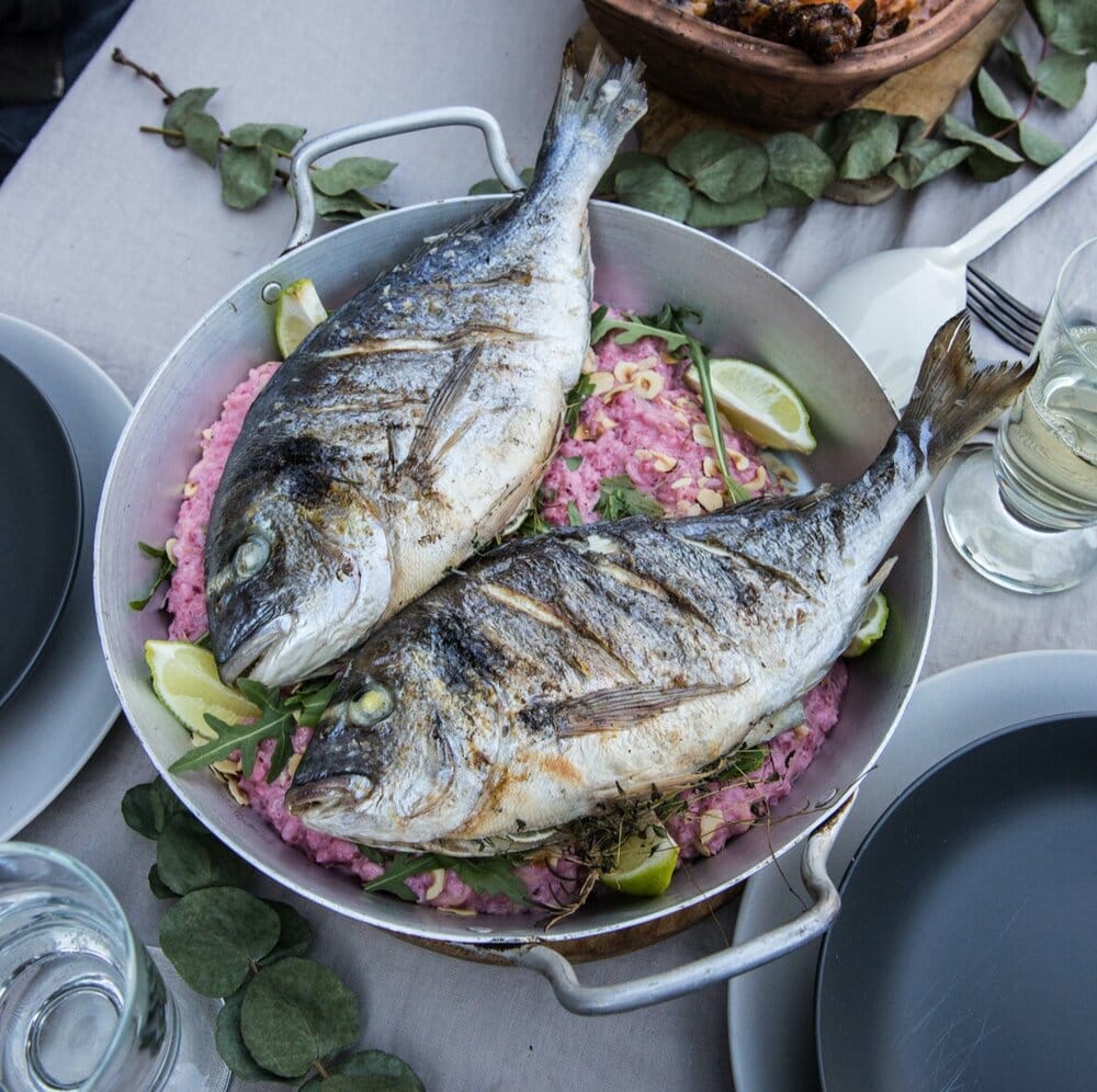 Zwei ganze gegrillte Doraden, serviert auf aromatischem Reis mit Limettenspalten in einer Metallpfanne auf einem gedeckten Tisch.