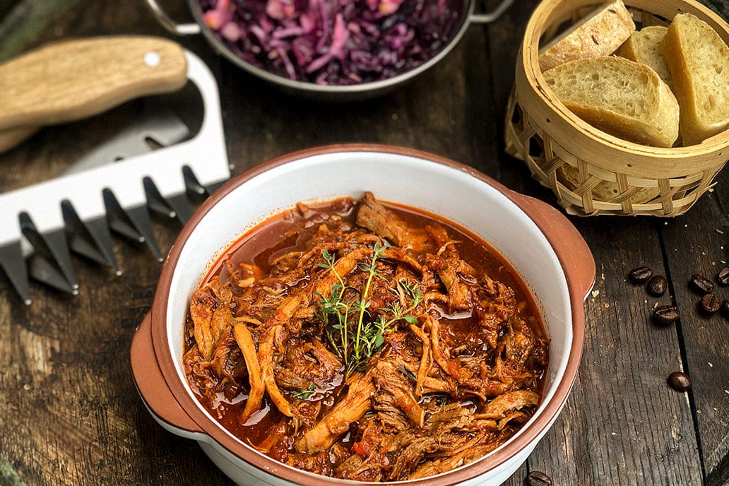 Zart geschmortes Pulled Pork in würziger Sauce, serviert mit frischem Brot und Rotkraut auf rustikalem Holztisch.