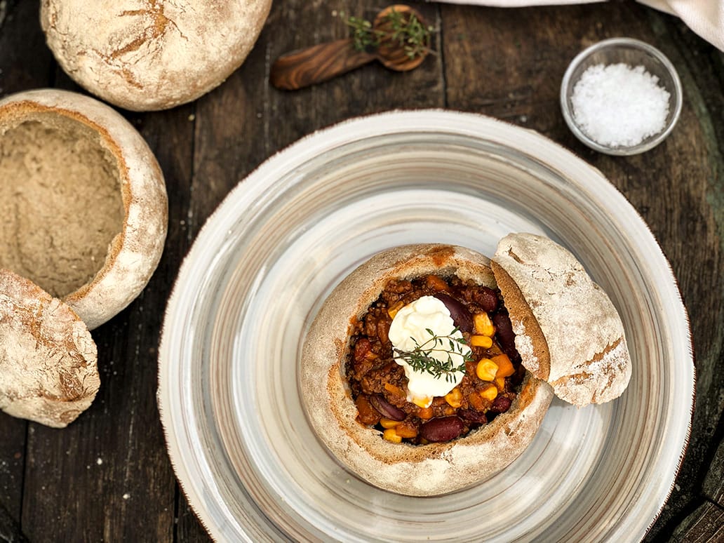 Chili con Carne aus dem Brottöpfchen Herzhaftes Chili con Carne, angerichtet im ausgehöhlten Brottopf, serviert mit Sauerrahm und frischen Kräutern.