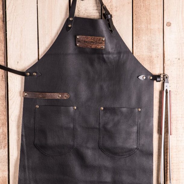 „Lederschürze fürs BBQ – Flatlay mit Grillzange | MERKLINGER“ Dateiname: merklinger-lederschuerze-flatlay-holz-grillzange.jpg MERKLINGER Lederschürze flach auf Holzuntergrund – zwei Taschen, Werkzeugleiste, Grillzange seitlich eingehängt.“