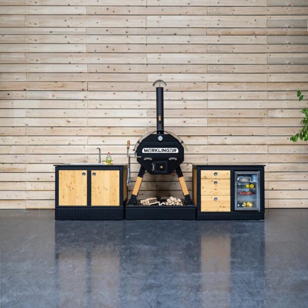Outdoor-Küchenzeile mit MERKLINGER Holzofen, Untertischkühlschrank und Spülmodul vor Hauswand.“