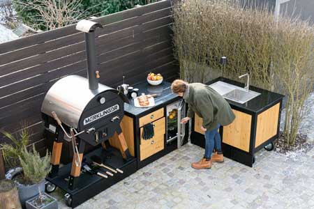 Luftaufnahme: Outdoor-Küche mit MERKLINGER Ofen; Person öffnet Glas-Kühlschrank zwischen Arbeits- und Spülmodul