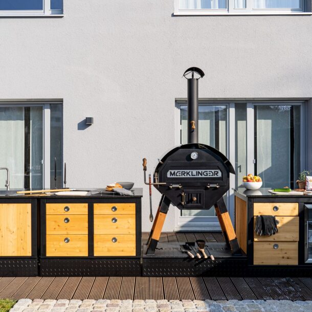 „MERKLINGER Outdoor-Küche auf der Terrasse mit Ofen, Stauraum und Kühlschrankmodul vor moderner Hausfassade.“