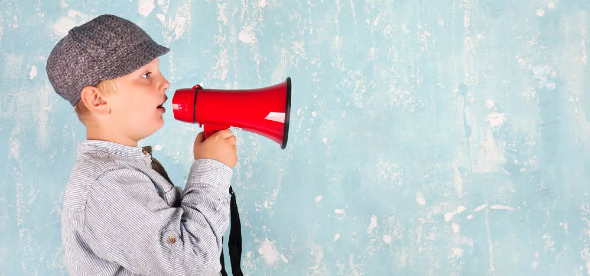 Junge mit Schiebermütze hält ein rotes Megafon an den Mund und ruft nach rechts; Hintergrund ist eine hellblaue, abgeplatzte Wand mit viel Freiraum für Text