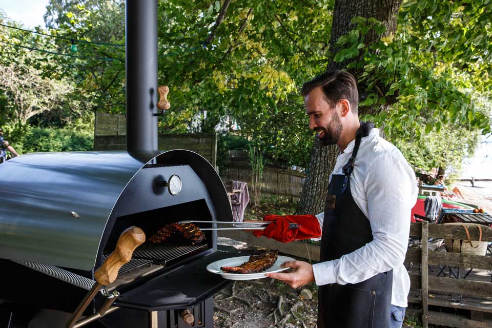Mann entnimmt Rippchen aus Rundofen Mann in Schürze entnimmt gegrillte Rippchen mit Zange aus MERKLINGER Rundofen im Garten