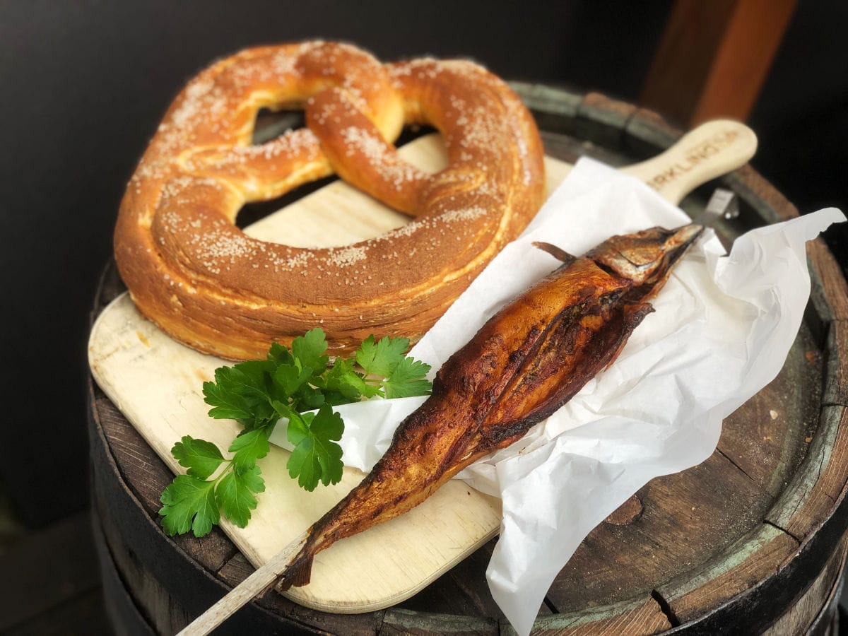 Frisch gebackene, goldbraune Riesenbrezel und geräucherter Fisch auf einem Holzbrett, dekoriert mit Petersilie.