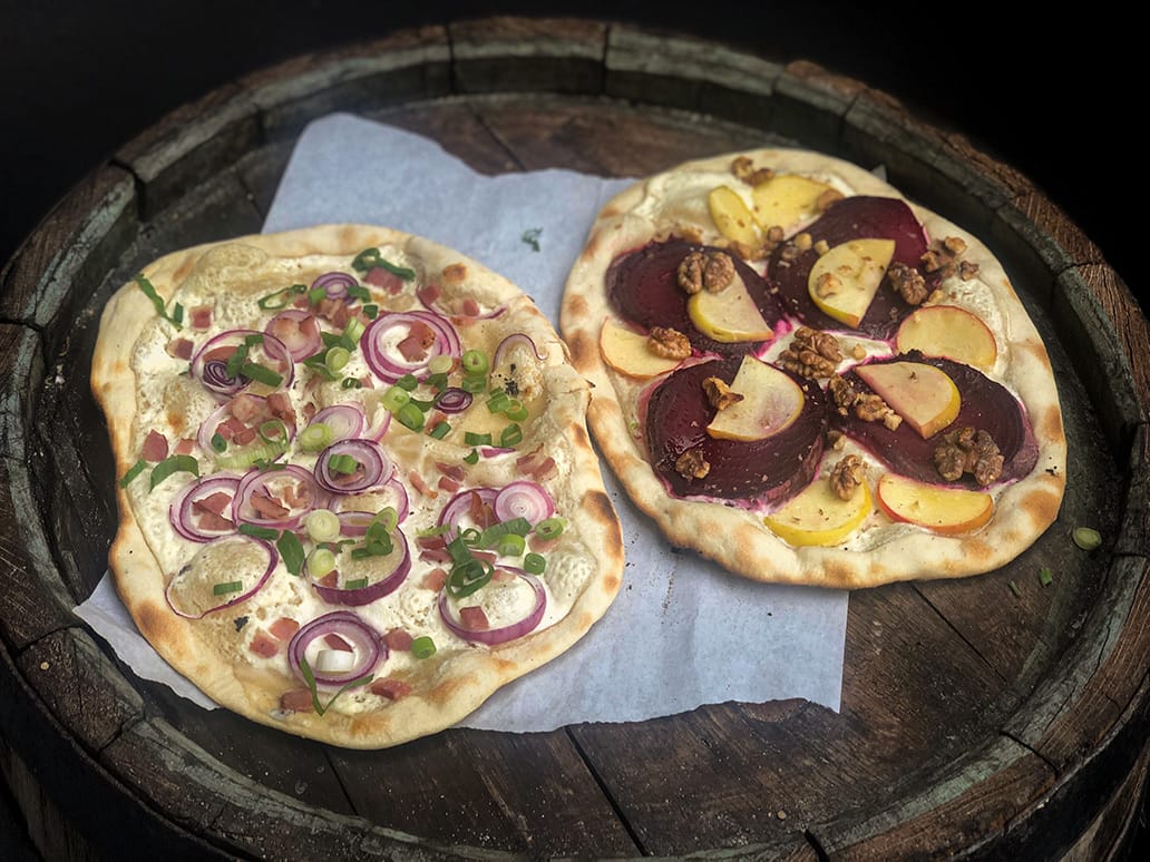 Zwei Flammkuchen auf Holzfass: klassisch mit Speck und Zwiebeln sowie vegetarisch mit roter Bete und Walnüssen, angerichtet auf Backpapier
