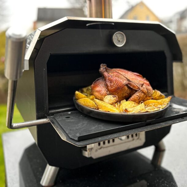 Ein kompakter, schwarzer Outdoor-Backofen mit geöffneter Klappe. Auf einer runden, dunklen Backplatte gart ein ganzes Brathähnchen, umringt von knusprigen Kartoffelspalten; im Hintergrund ein Garten/Hausszenerie. Ein kompakter, schwarzer Outdoor-Backofen mit geöffneter Klappe. Auf einer runden, dunklen Backplatte gart ein ganzes Brathähnchen, umringt von knusprigen Kartoffelspalten; im Hintergrund ein Garten/Hausszenerie Merklinger Bobby Keltenschale