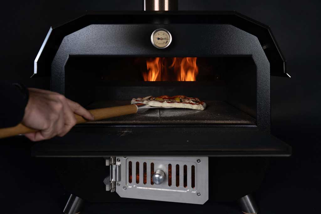 Pizza backt auf Steinplatte im MERKLINGER Holzbackofen vor lodernder Flamme.