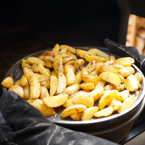 Kartoffelwedges auf der Platte: Goldbraun gebackene Kartoffelspalten auf runder, schwarzer Keramik-Backplatte, gehalten mit Hitzeschutzhandschuhen.